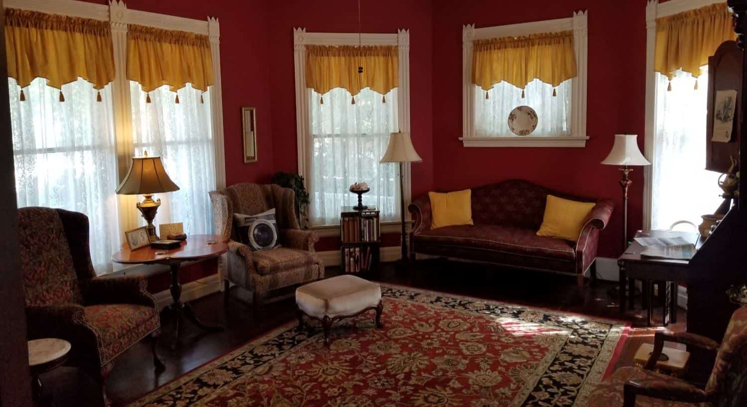 Large room with burgundy walls, dark hardwood floors, area rug, Victorian chairs and sofa, and many large windows