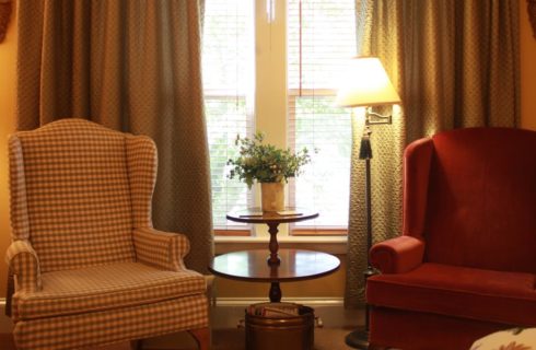 Sitting area in bedroom with plaid arm chair, solid burgundy arm chair, and wooden table near large windows