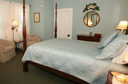Bedroom with blue walls, dark carpeting, four-post wooden bed with blue bedding, wooden dresser, and small sitting area