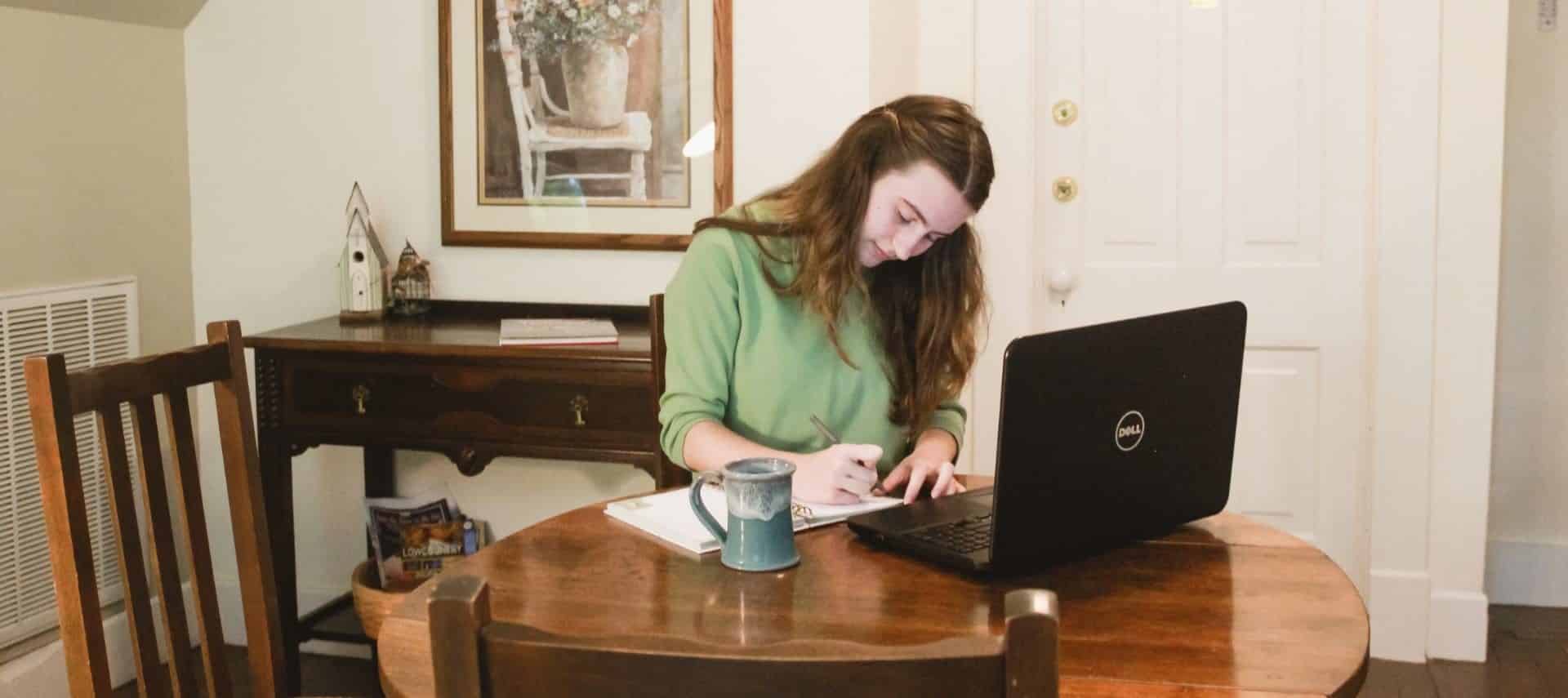 Woman in green shirt sitting at desk writing with laptop near by