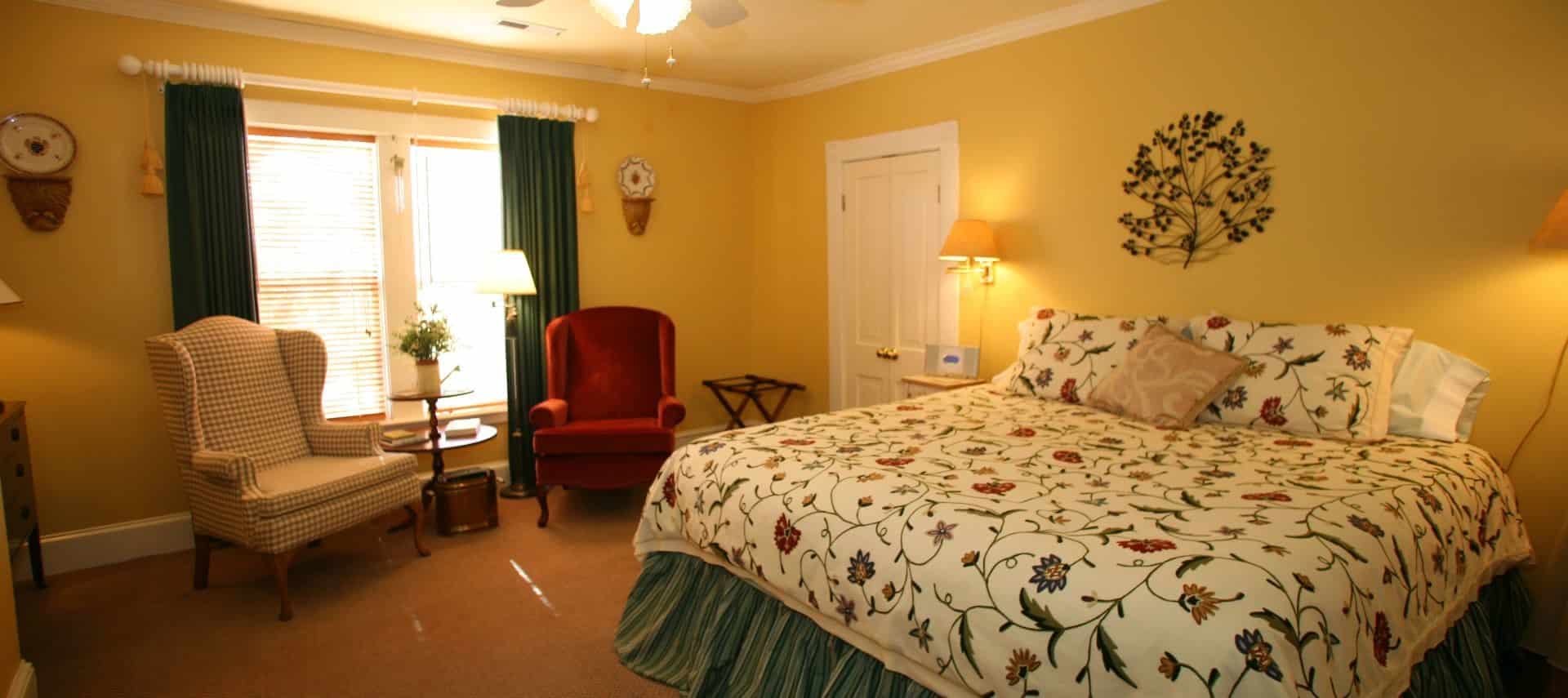 Bedroom with yellow walls, carpeting, floral bedding with green bed skirt, and sitting area with arm chairs and small table