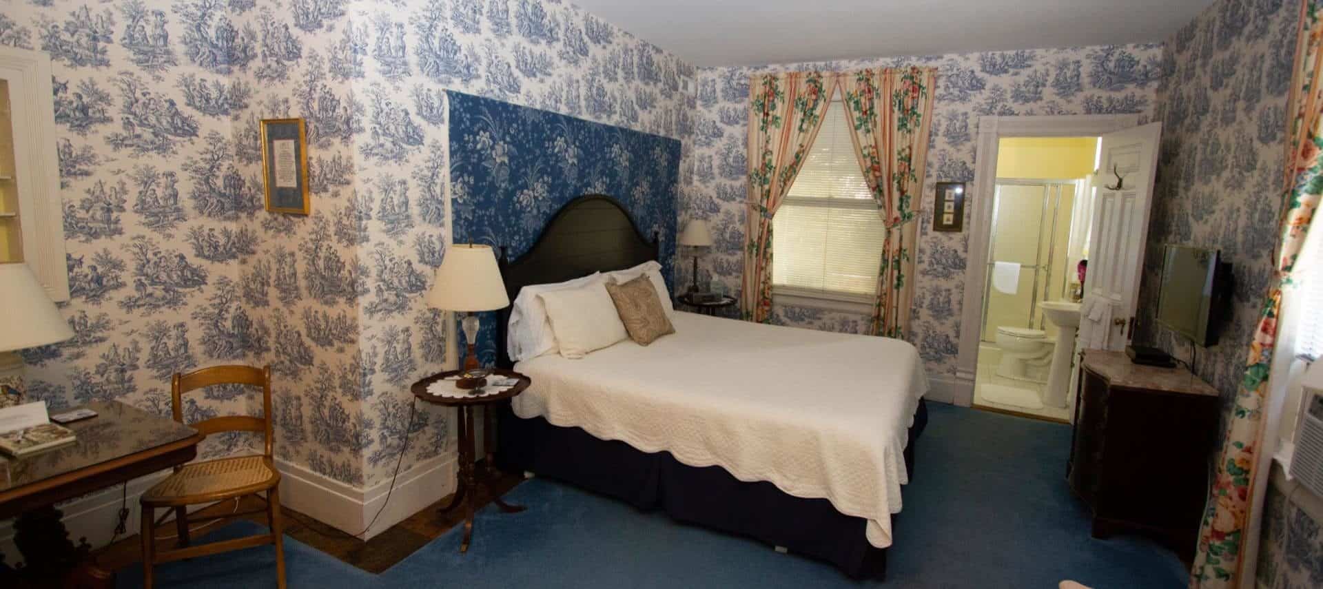 Bedroom with blue and white Victorian wall paper, blue carpet, dark wood bed with white linens, and view into the bathroom